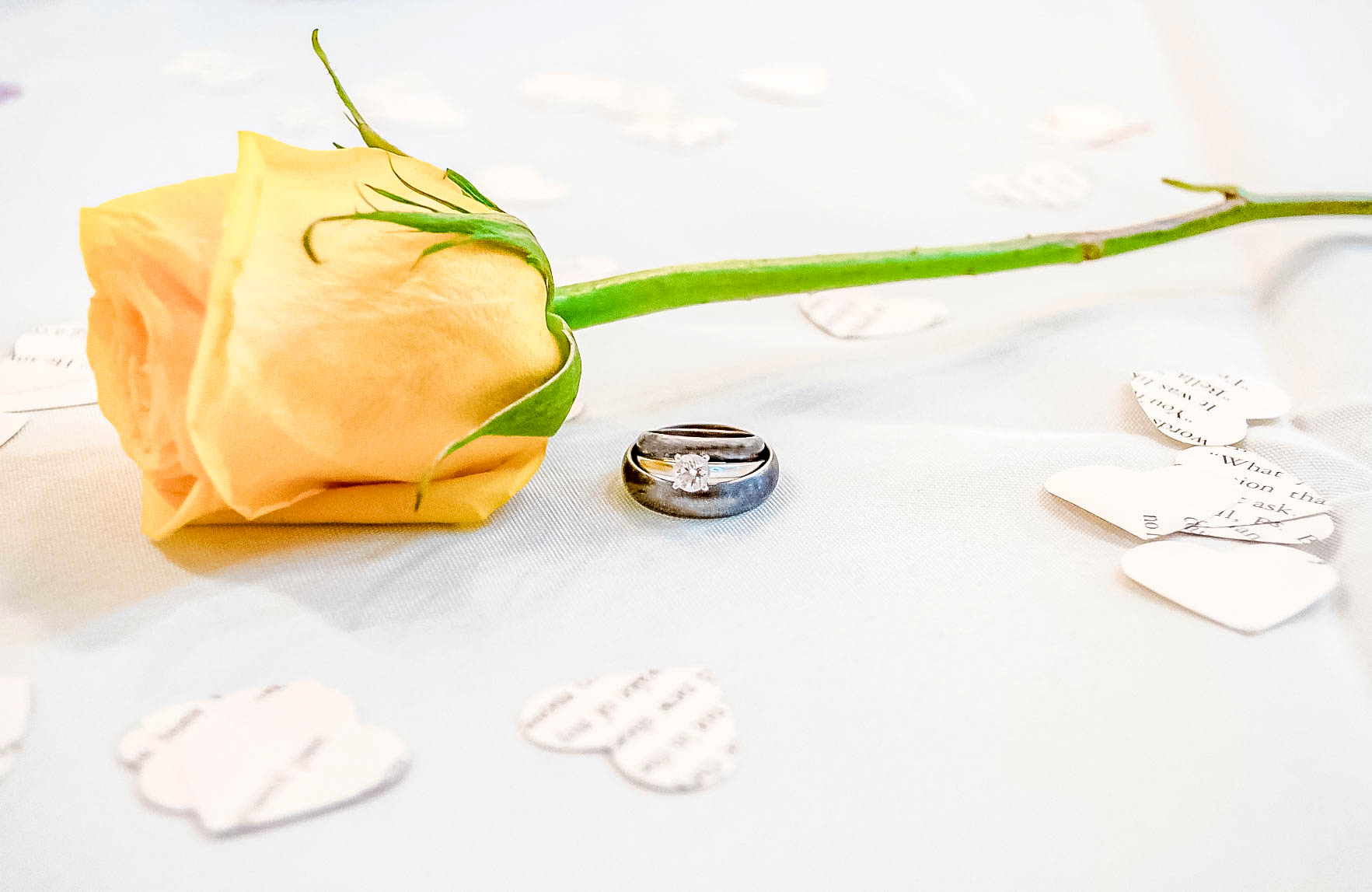 wedding rings stacked on table by yellow long stem rose and paper heart confetti
