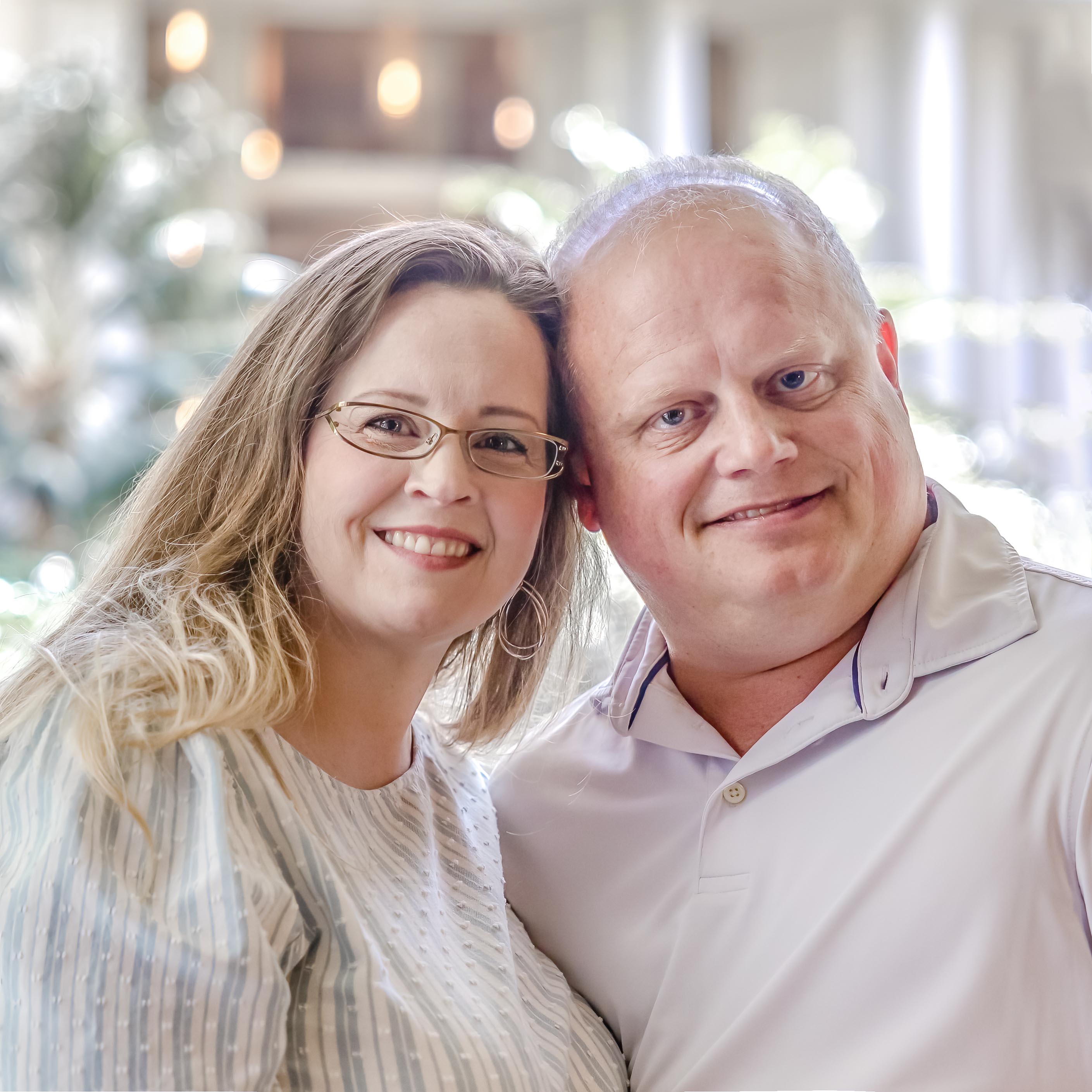 A close-up portrait of Matt and Riley-Payton, the husband and wife team behind Bella Lumiere Photography, smiling together in a softly lit, romantic setting. The blurred background with warm bokeh lighting enhances the light and airy aesthetic of their wedding photography brand.