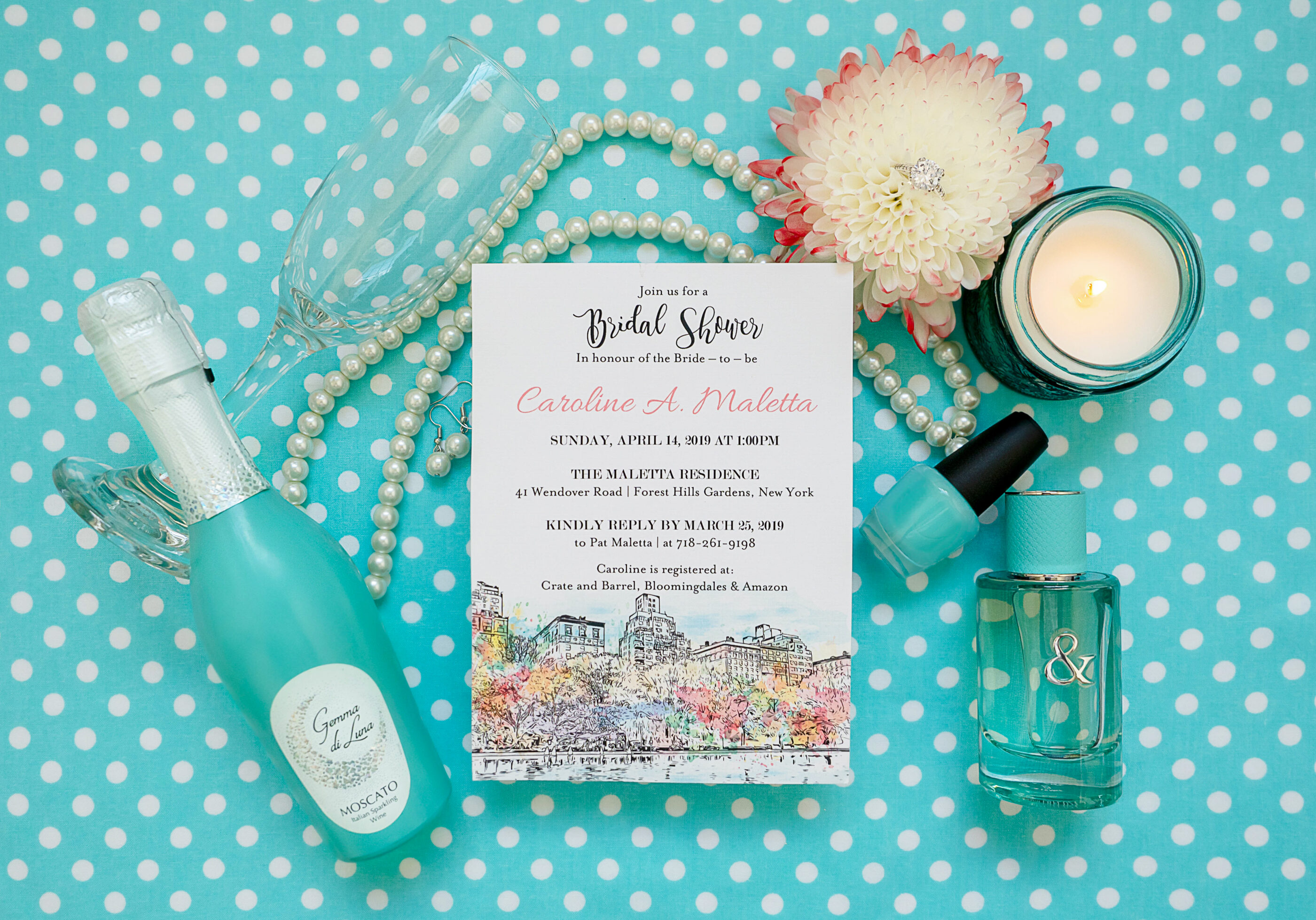 Flat lay of a bridal shower invitation on a teal polka dot background, surrounded by pearls, a flower with a ring in the center, champagne glasses, a bottle of Moscato, teal perfume, nail polish, and a candle.