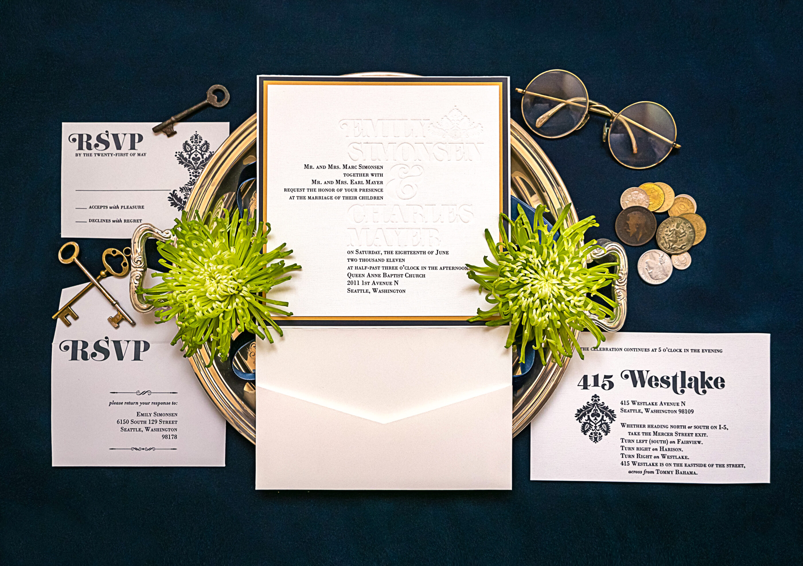 Flat lay of a formal wedding invitation suite with embossed text on a navy background, accented with antique keys, vintage coins, green spider mums, a silver tray, and gold-rimmed glasses for an elegant Seattle wedding.