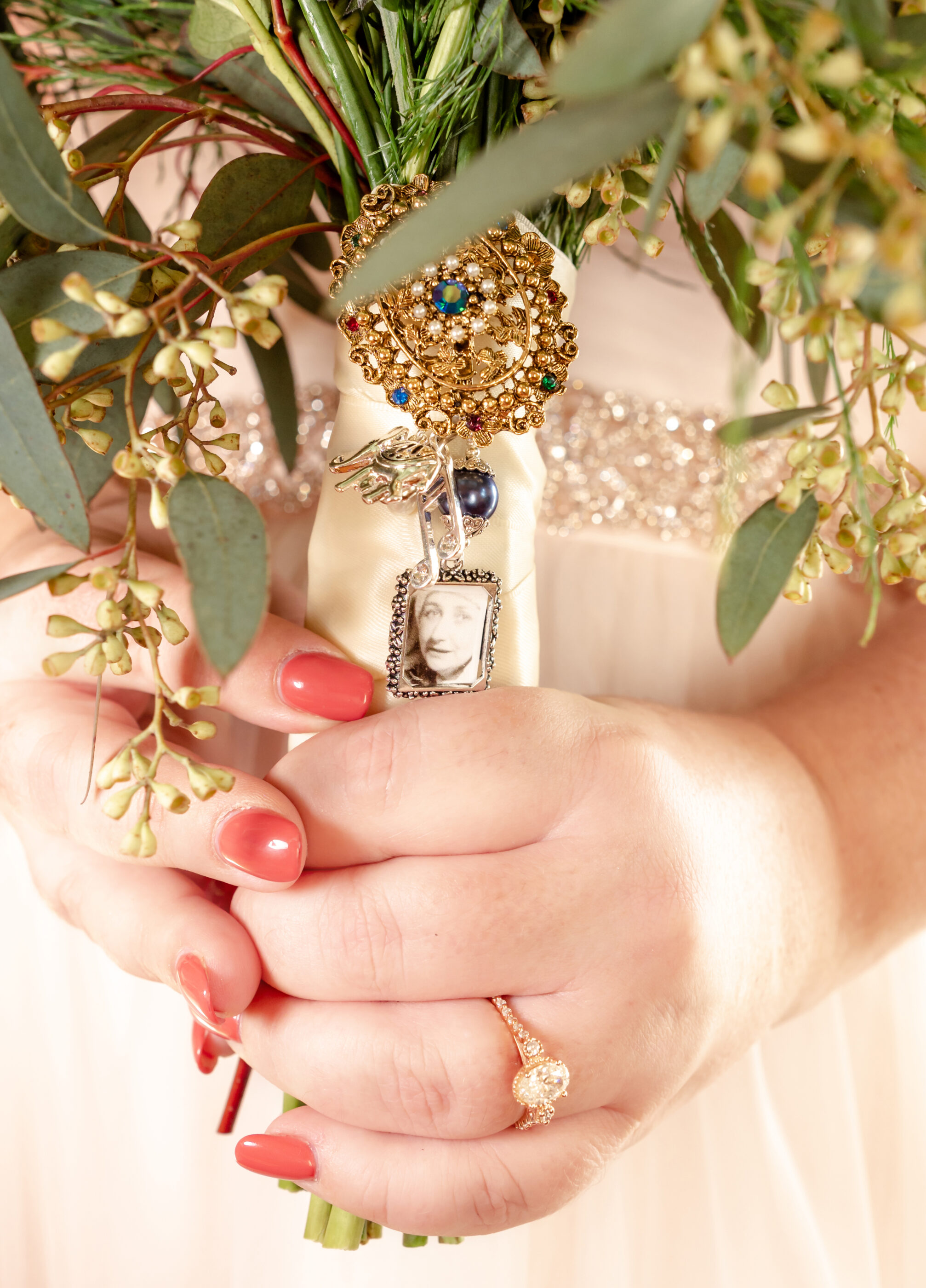 Close-up of bride’s hands holding bouquet with gold brooch and photo charm honoring a loved one.