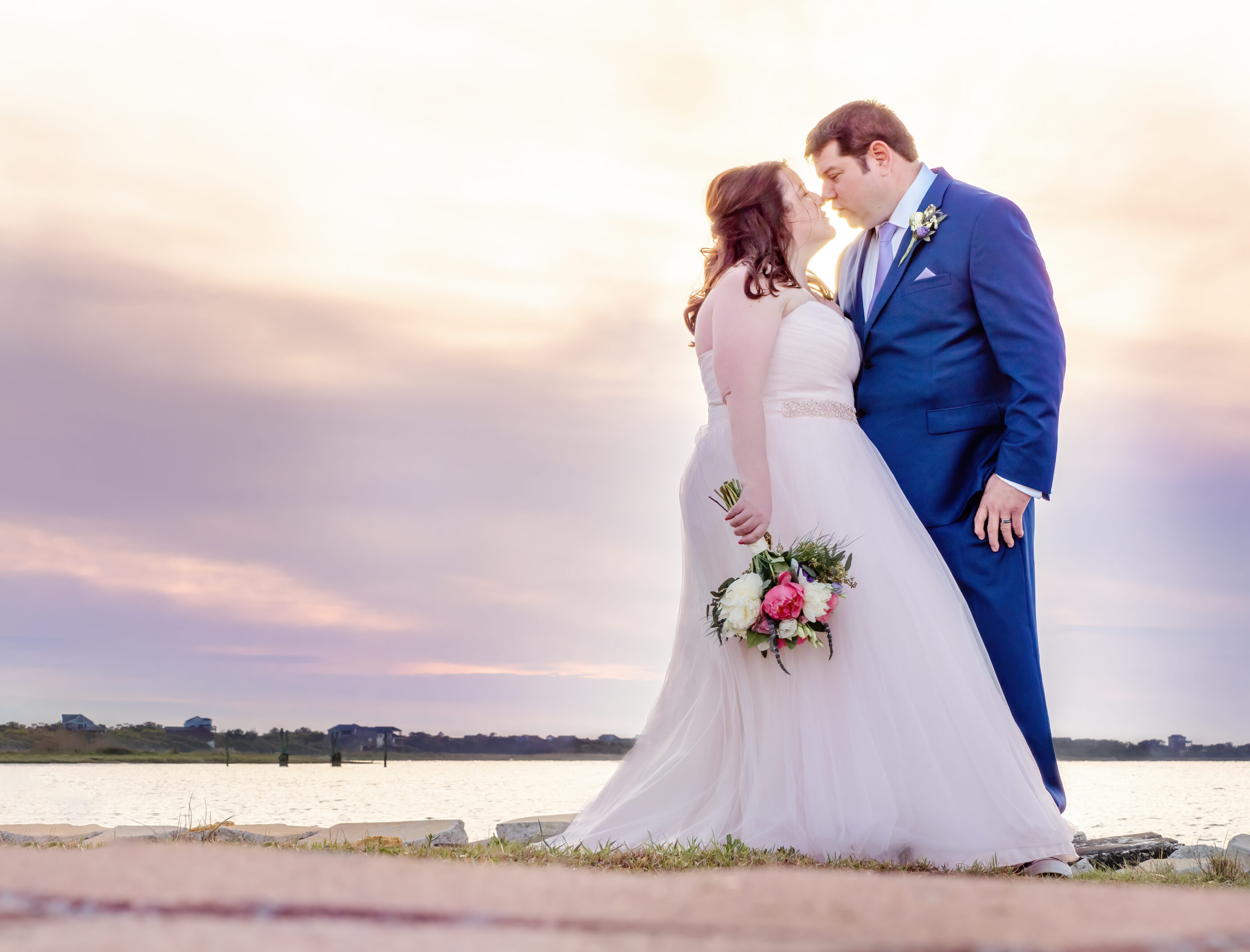 Bride and groom share a romantic kiss by the water at sunset, bride holding bouquet.