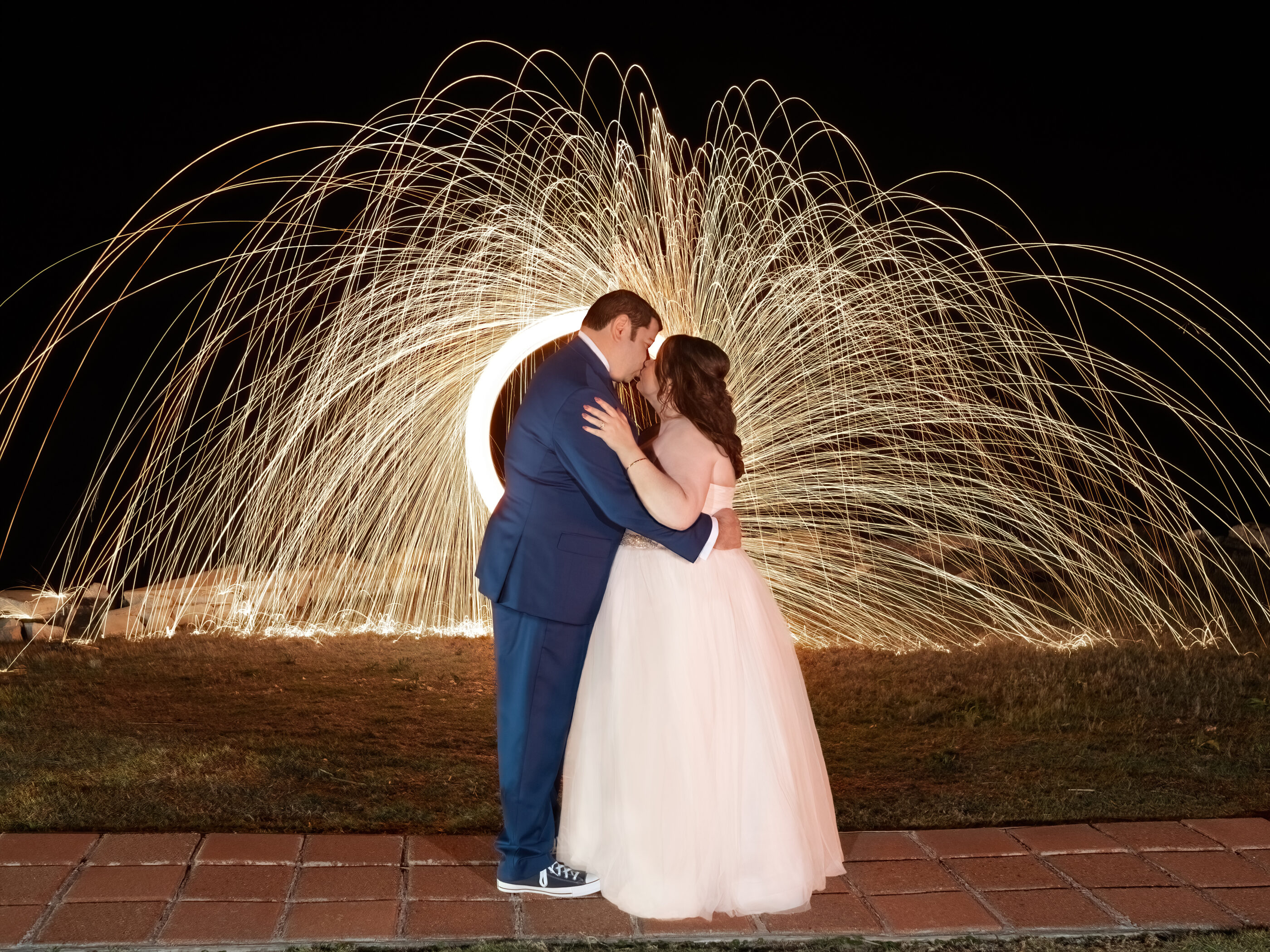 Bride and groom kiss at night with steel wool sparkler effect creating dramatic light trails behind them during wedding photography and videography.