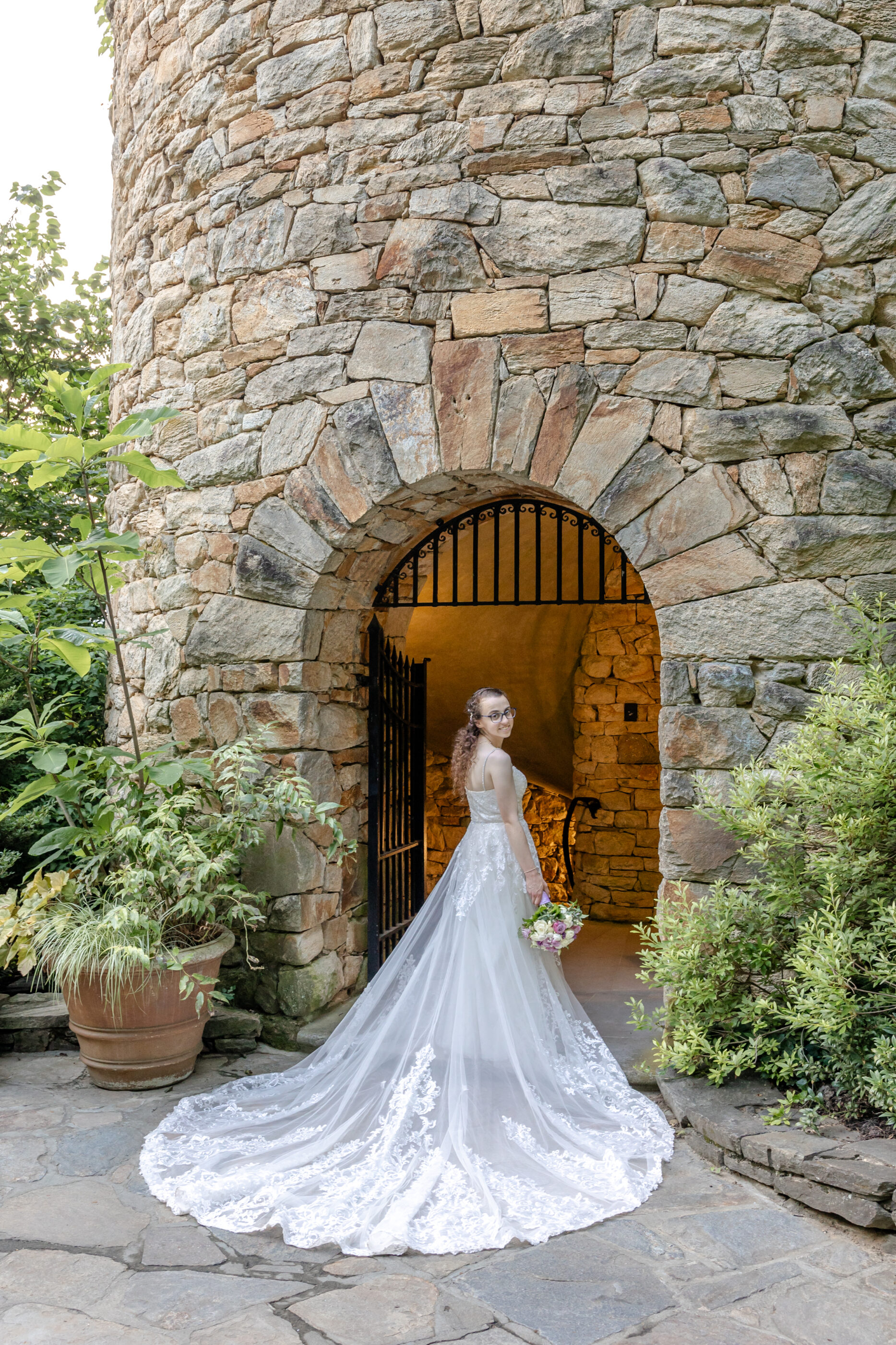 Bride in lace wedding gown stands in a stone archway, looking over her shoulder while holding bouquet.