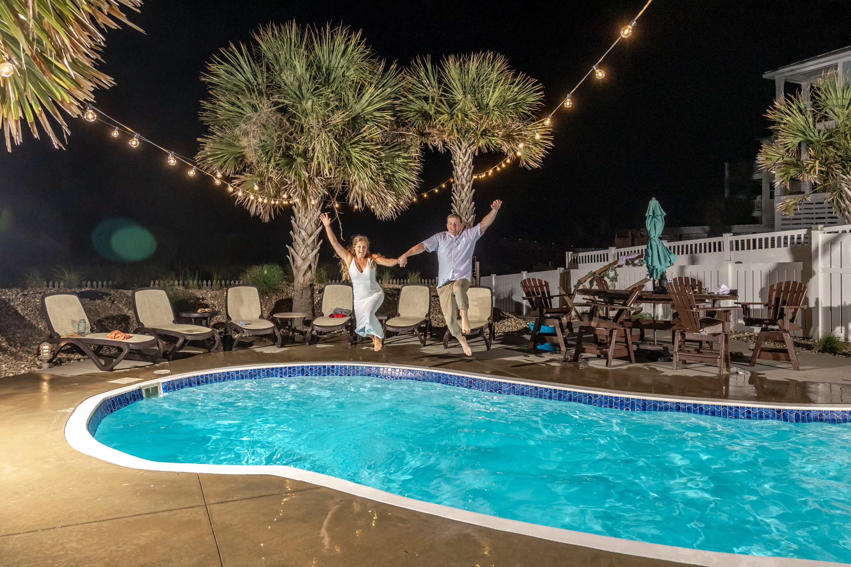 Bride and groom in midair holding hands while jumping into a pool at night.