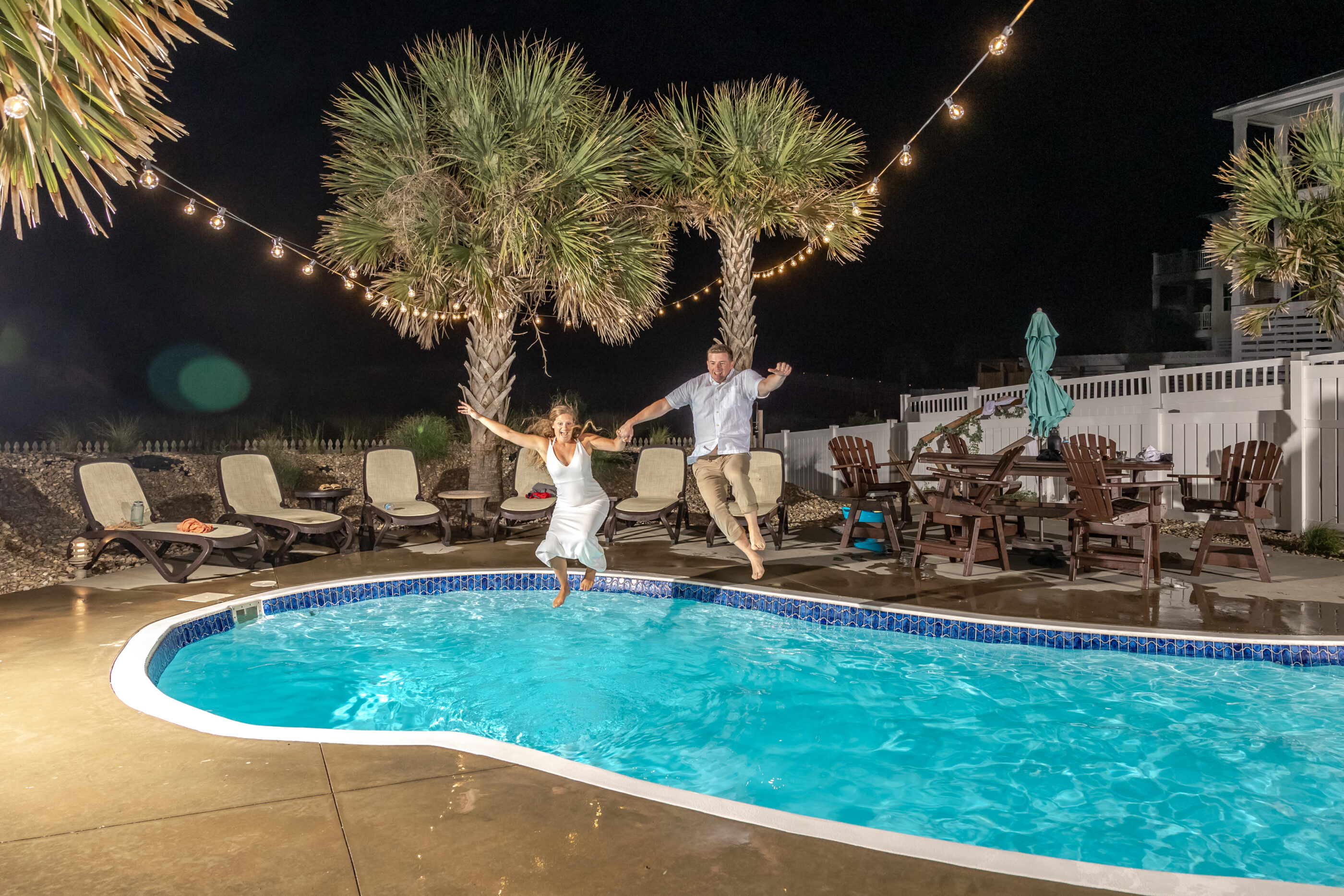 Bride and groom about to land in a pool together while holding hands during a nighttime celebration.