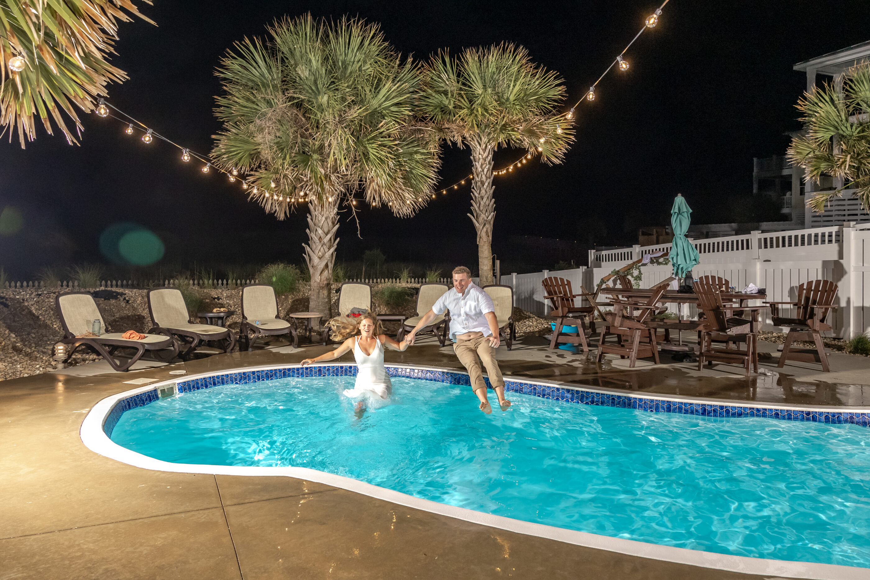 Bride lands in pool while groom is still midair, both laughing and holding hands.