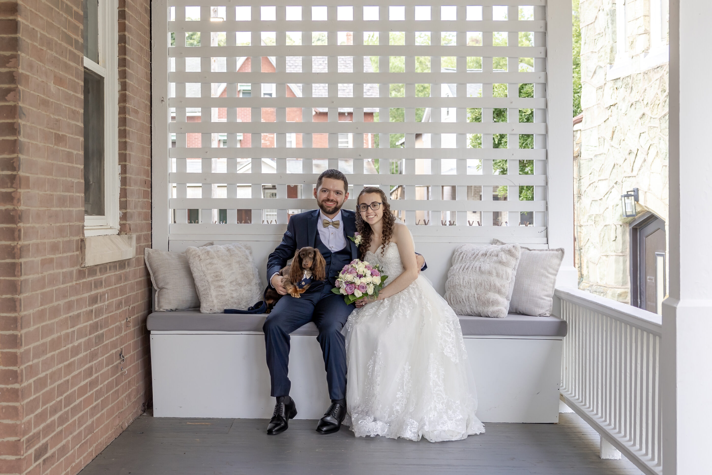 Bride and groom sit on porch bench with their small brown dog, smiling and holding bouquet.