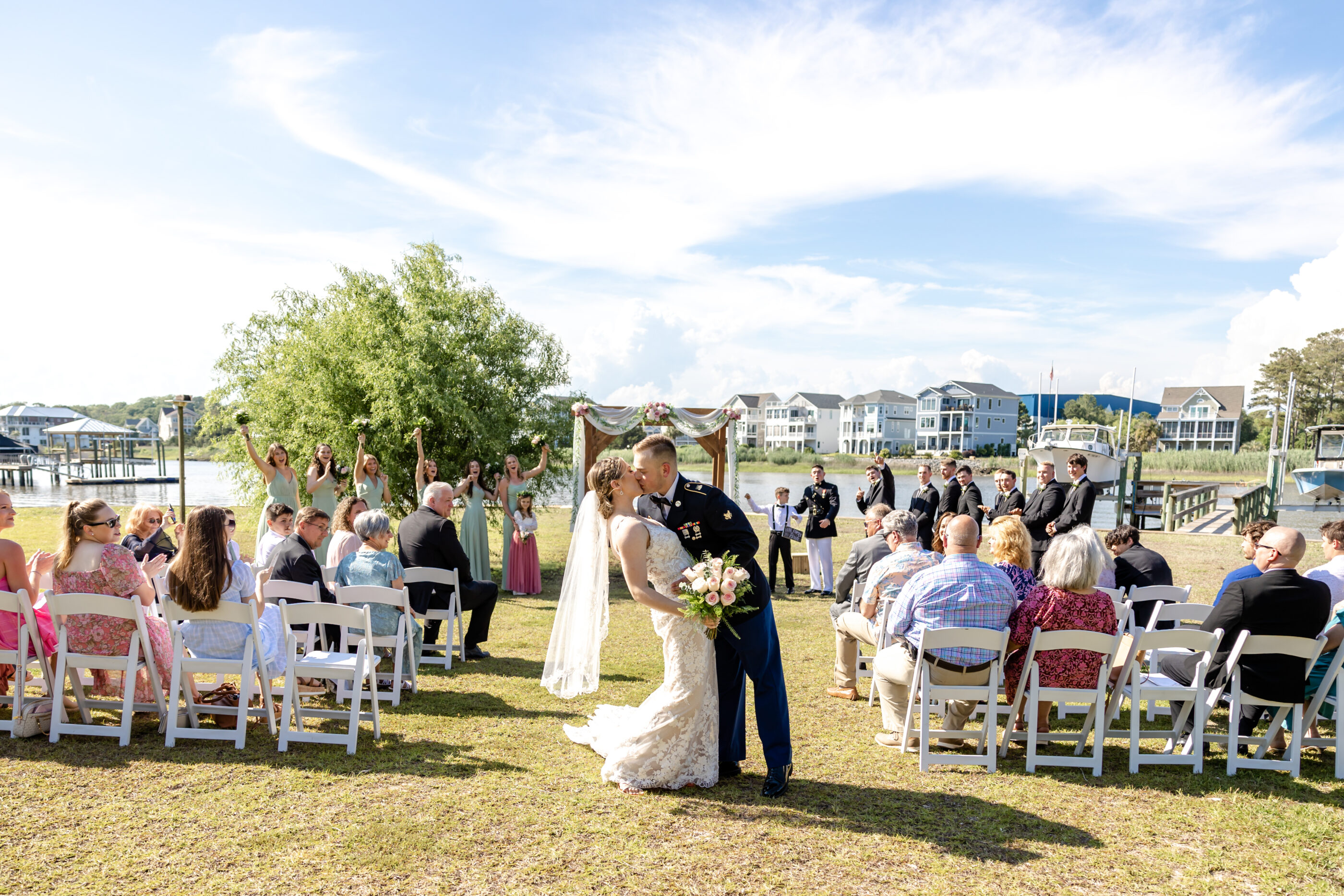 Bride and groom share first kiss under ceremony arch at a waterfront while wedding photographer and videographer capture moment and guests and bridal party cheer.