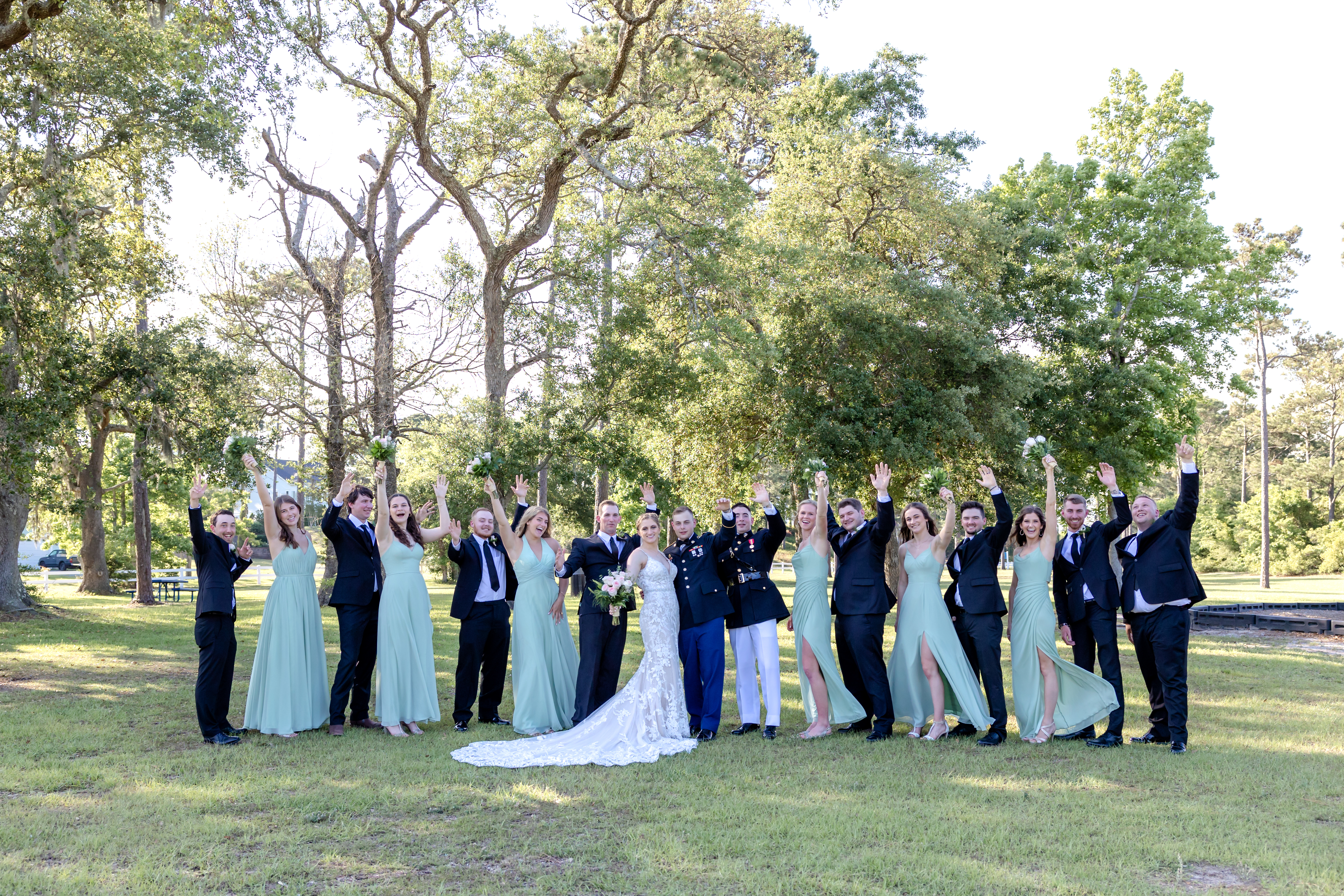 Wedding party cheers with arms raised as bride and groom stand together in a wooded park.