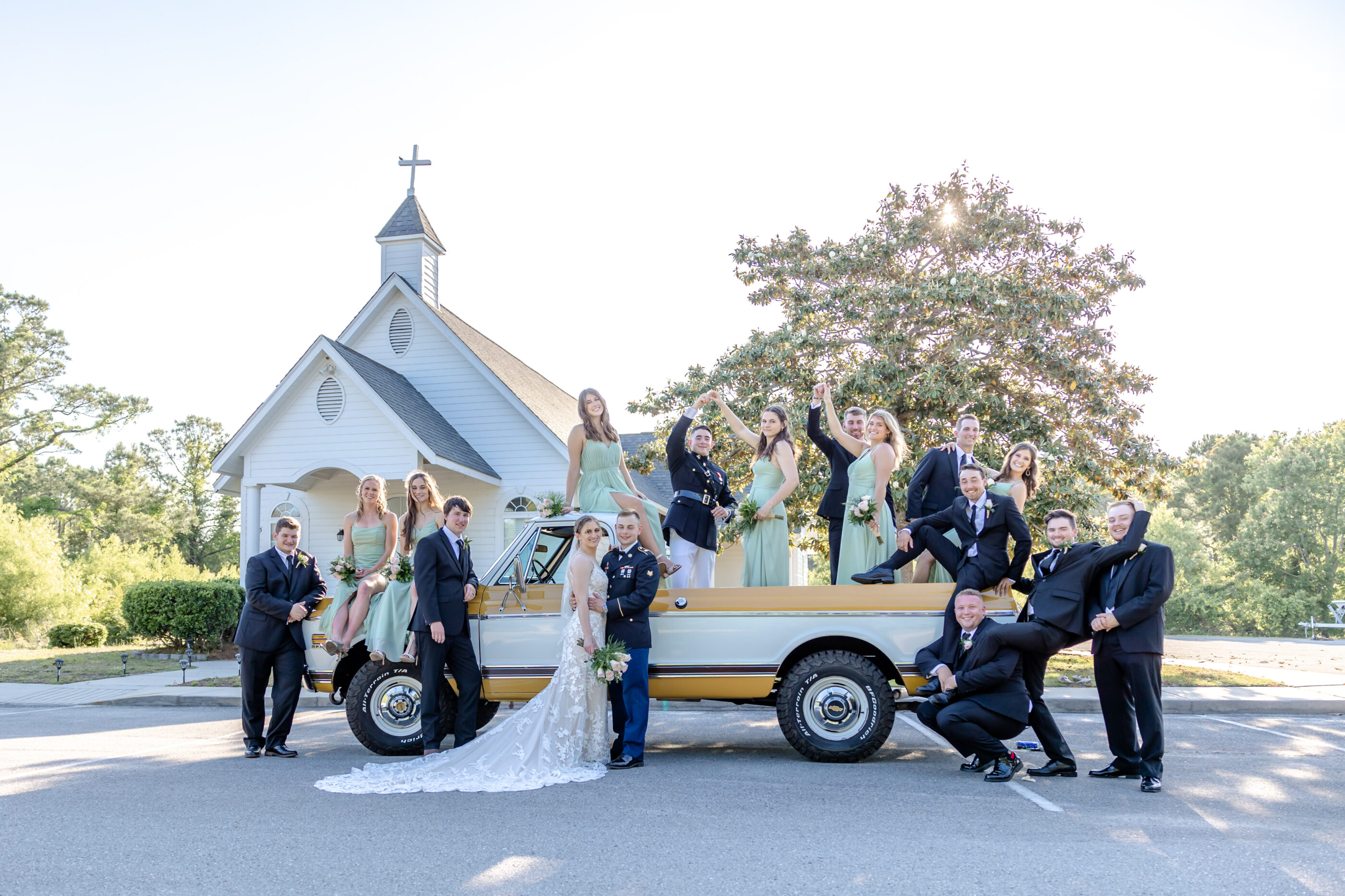 Bride, groom, and wedding party pose creatively on and around a vintage yellow truck parked in front of a white chapel.